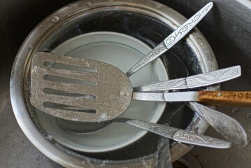 a pile of dirty dishes from bowls, plates and metal forks with spoons in water in a gray metal sink...