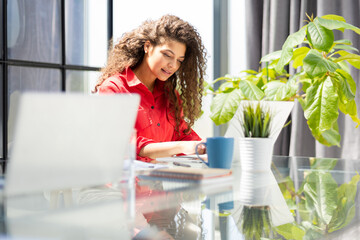 Attractive cheerful business woman in red shirt working on laptop at modern office.