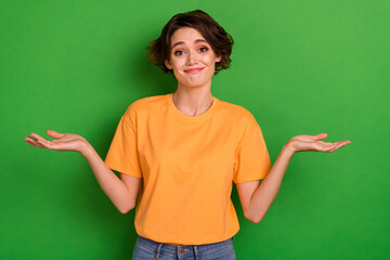 Photo of doubtful funny young woman dressed orange t-shirt shrugging shoulders isolated green color...