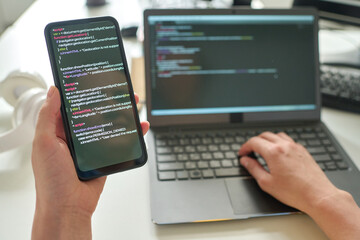 Close-up of programmer holding smartphone with codes in her hands and connecting it with laptop at...