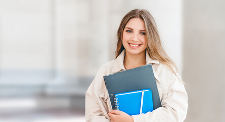 Young student girl smiling against university. Cute girl student holds folders and notebooks in...