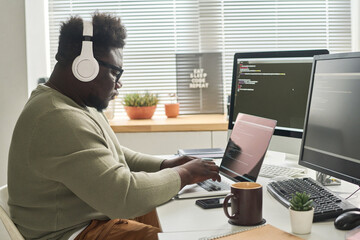 African IT programmer in wireless headphone concentrating on his online work on laptop, he writing computer codes to program - obrazy, fototapety, plakaty