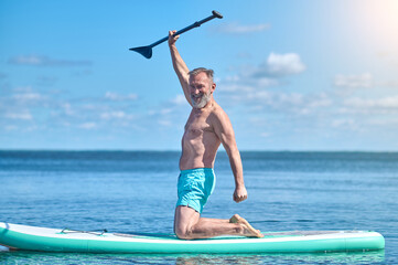 Man lifting paddle smiling at camera kneeling on surfboard