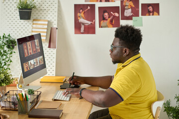 African retoucher sitting at his workplace and using graphic tablet to retouch photo on computer...
