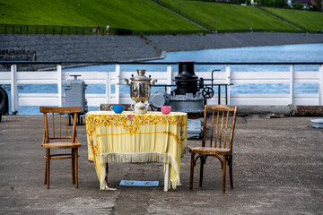 a table with a tablecloth and a samovar on a platform at the pier of ships on a summer day