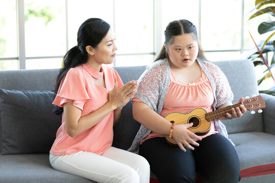mother teaching and encourage to a girl with down syndrome singing and playing ukulele or small guitar on sofa