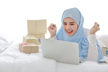 Beautiful and young asian woman in muslim sleepwear with attractive look, lies on bed with computer, mobile phone and online package box delivery. Smart girl with hijab receive good news and surprise.