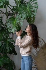Being in touch with nature. Young attractive German woman plant lover caring for plants, standing near big Monstera houseplant and touching leaf, enjoying leisure time by doing home gardening