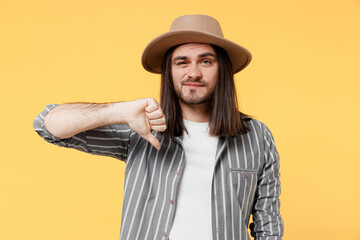 Young sad dissatisfied man he 20s wearing striped grey shirt white t-shirt hat showing thumb down dislike gesture isolated on plain yellow color background studio portrait. People lifestyle concept.