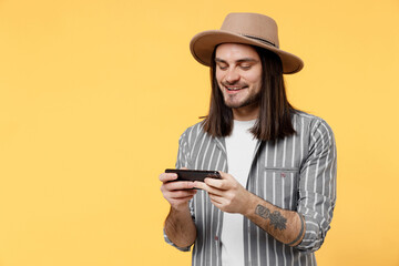 Gambling young man he wears striped grey shirt white t-shirt hat using play racing app on mobile cell phone hold gadget smartphone for pc video games isolated on plain yellow color background studio