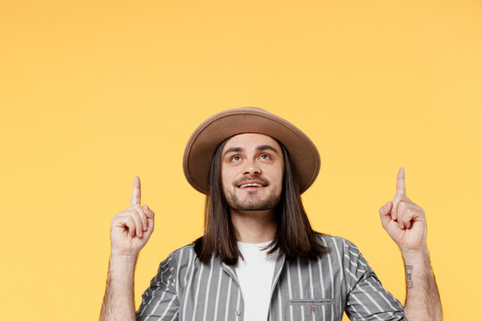 Young fun man he 20s wears striped grey shirt white t-shirt hat pointing index finger indicate overhead on workspace area copy space mock up isolated on plain yellow color background studio portrait