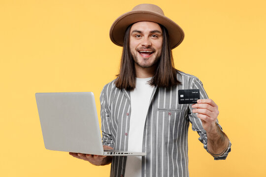 Young man he 20s wears striped grey shirt white t-shirt hat using laptop pc computer hold credit bank card doing online shopping order delivery booking tour isolated on plain yellow color background.