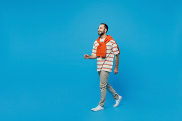 Full body side profile view young smiling caucasian cheerful man 20s wearing orange striped t-shirt walking going strolling isolated on plain blue background studio portrait. People lifestyle concept.