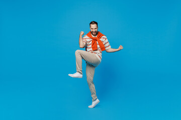 Fototapeta na wymiar Full body young excited man 20s in orange striped t-shirt doing winner gesture celebrate clench fists say yes raise up leg isolated on plain blue background studio portrait. People lifestyle concept.