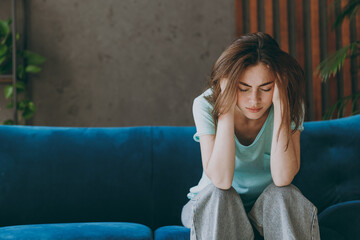 Young frustrated ill woman she 20s wear casual clothes mint t-shirt holding head suffer from headache close eyes sit on blue sofa indoor rest at home in own room apartment. People lifestyle concept - obrazy, fototapety, plakaty