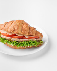 Fresh croissant sandwich with chicken, tomatoes and salad leaf on white plate, isolated. Breakfast in french. Side view.       