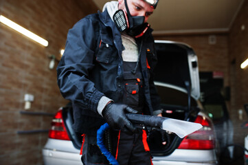 Man in uniform and respirator, worker of car wash center, cleaning car interior with hot steam cleaner. Car detailing concept.