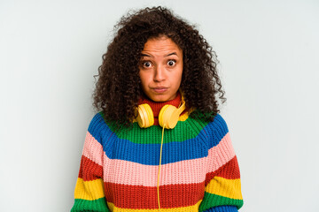 Young Brazilian woman listening to music isolated on blue background shrugs shoulders and open eyes...