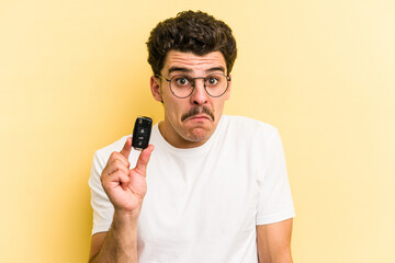 Young caucasian man holding car keys isolated on yellow background shrugs shoulders and open eyes confused. - obrazy, fototapety, plakaty