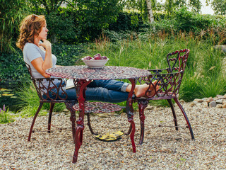 A pensive brunette in her 50s sits cross-legged, barefoot, on a chair in her garden, enjoying the silence and fresh fruit. Healthy calm lifestyle, vegetarian food.