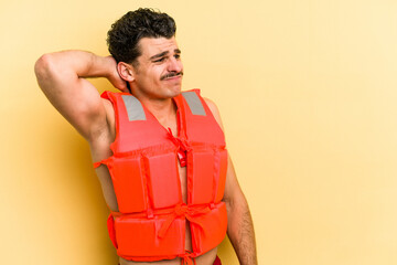 Young caucasian man wearing life jacket isolated on yellow background touching back of head,...