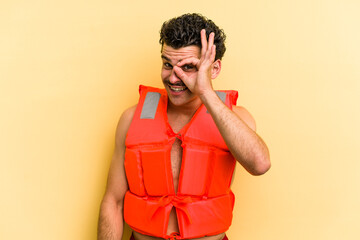 Young caucasian man wearing life jacket isolated on yellow background excited keeping ok gesture on...