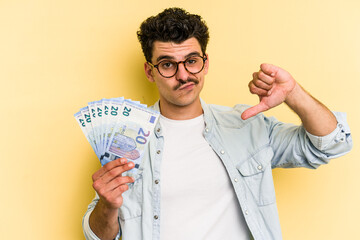 Young caucasian man holding banknotes isolated on yellow background showing a dislike gesture,...