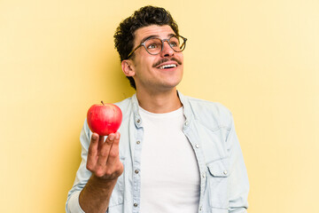 Young caucasian man holding an apple isolated on yellow background dreaming of achieving goals and...