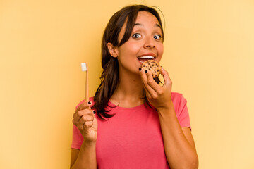 Young hispanic woman washing teeth after eating cookies isolated on yellow background