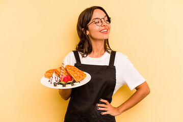 Young hispanic dependent woman holding waffles isolated on yellow background looks aside smiling, cheerful and pleasant.