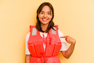 Young hispanic woman holding life jacket isolated on yellow background person pointing by hand to a...