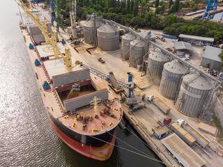 Loading grain into sea cargo vessel in seaport from silos of grain storage. Aerial view