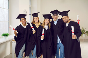 Portrait of smiling diverse students in mantles and hats pose with diplomas celebrate university...