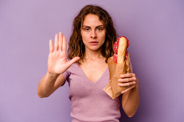 Young caucasian woman eating a sandwich isolated on purple background standing with outstretched hand showing stop sign, preventing you.