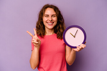 Young caucasian woman holding a clock isolated on yellow background showing number two with fingers.