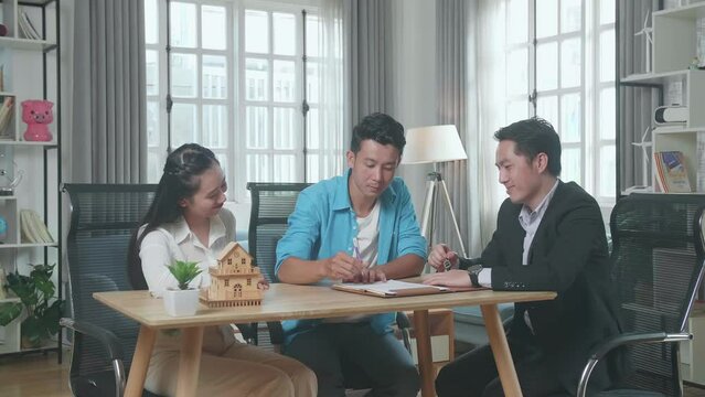 Asian Man Sitting With A Woman Receiving The Keys From A Real Estate Agent After Signing On House Purchase Contract Paper
