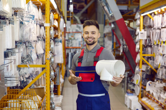 Portrait of happy shop assistant who works at DIY retail store. Cheerful handsome man in uniform standing in aisle, showing white PVC T shaped ventilation pipe joint, looking at camera and smiling