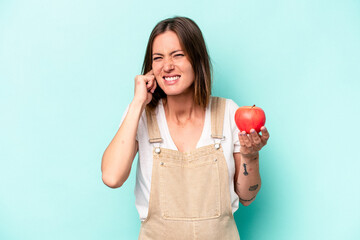 Young caucasian pregnant woman holding an apple isolated on blue background covering ears with hands.