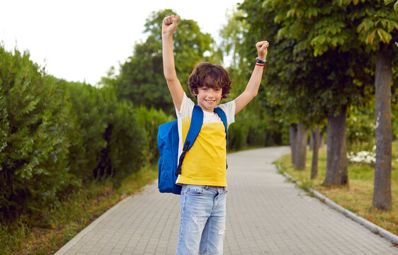 Little child happy to go back to school. Cute student boy in yellow white T shirt with blue bag standing on path in green park, raising his hands up, looking at camera and smiling. School, fun concept