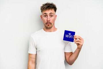 Young caucasian man holding wifi placard isolated on white background shrugs shoulders and open eyes confused.