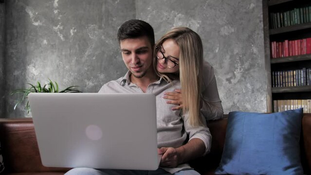 young guy working on laptop at home when girlfriend coming to him and hugging and smile