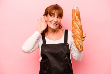 Middle age baker woman isolated on pink background trying to listening a gossip.
