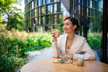 Young business woman holding and drinking coffee in street cafe in city. Portrait of beautiful woman worker waiting client on street of Prague