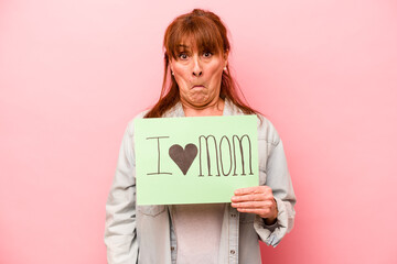 Middle age caucasian woman holding I love mom placard isolated on pink background shrugs shoulders and open eyes confused.