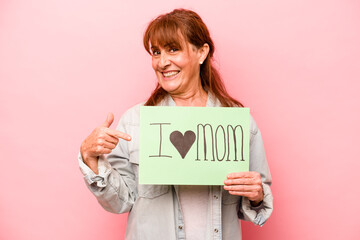 Fototapeta na wymiar Middle age caucasian woman holding I love mom placard isolated on pink background smiling and pointing aside, showing something at blank space.