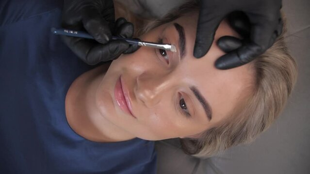 Master Beauty applies white markings to correct shape of eyebrows with a special brush. The beautiful face of young girl model on sofa in a beauty salon.Slow Motion.Top View
