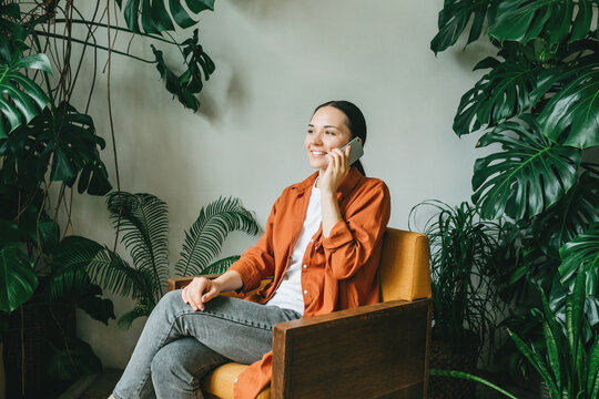 A young smiling woman is sitting at home in a yellow armchair in a green garden and talking on the phone.  The concept of remote work or training. The concept of eco-friendly housing and minimalism