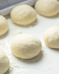 Fototapeta na wymiar Homemade pizza dough with wheat flour dusted on a white kitchen table. Freshly kneaded bread buns ready to be baked. Vertical shot. A closeup.