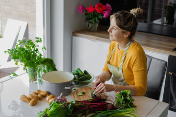 Glad and calm blonde woman in apron chopping aromatic vegetables, looking out window. Detox diet...