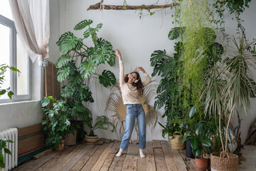Positive young italian woman in wireless headphones listening to music with pleasure, enjoying the moment, relaxing, dancing on wooden floor in cozy home garden with monstera and tropical plants.  - obrazy, fototapety, plakaty